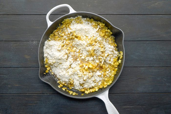 A white skillet on a dark wooden surface contains chopped yellow vegetables and a pile of flour, ready to be mixed together.