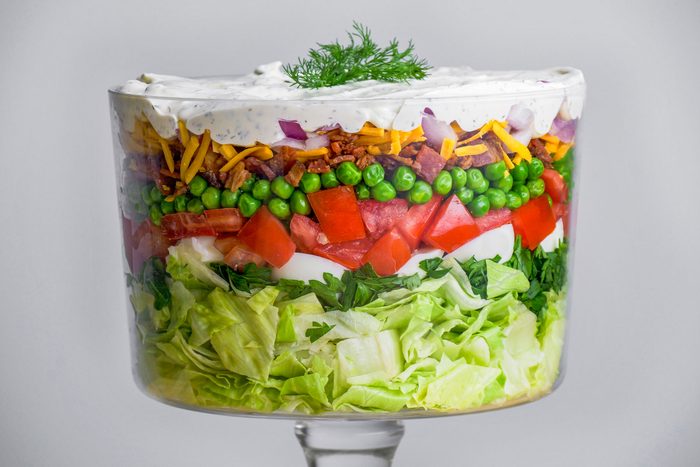 Close-up shot of a 7-Layer Salad in a clear trifle bowl, showing layers of lettuce, spinach, tomato, peas, cheese, bacon bits, onion, and creamy dressing;