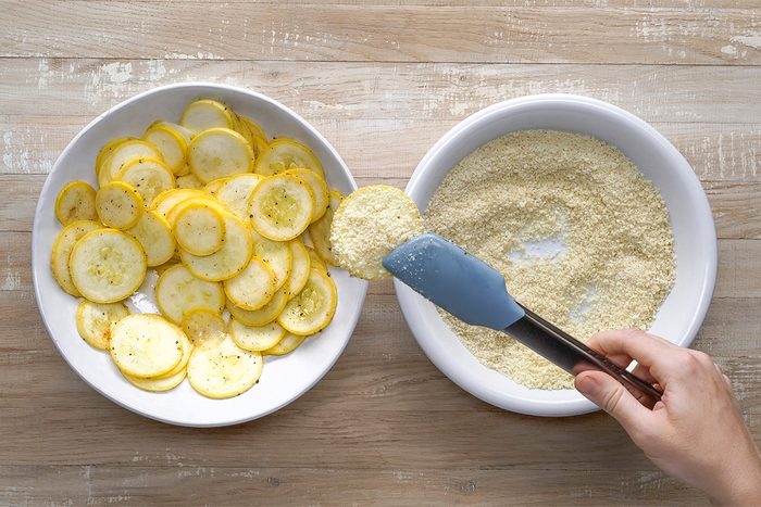 A hand uses tongs to dip a slice of yellow squash into a bowl of breading mixture, with a plate of sliced squash on the side, all on a wooden surface.