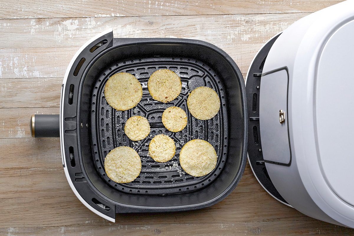 An air fryer basket containing eight round slices of food, possibly breaded vegetables or chips, arranged in a single layer on a wooden surface. The air fryer lid is partially removed, showing the contents inside.