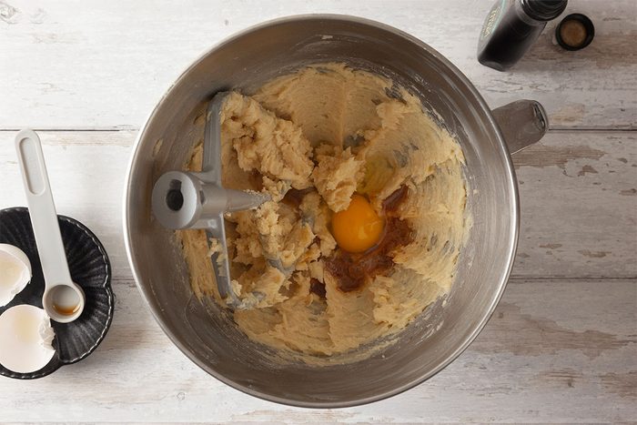 A mixing bowl with creamed butter and sugar, a cracked egg, and vanilla extract about to be mixed. Eggshells and a measuring spoon are nearby on a light wooden surface.
