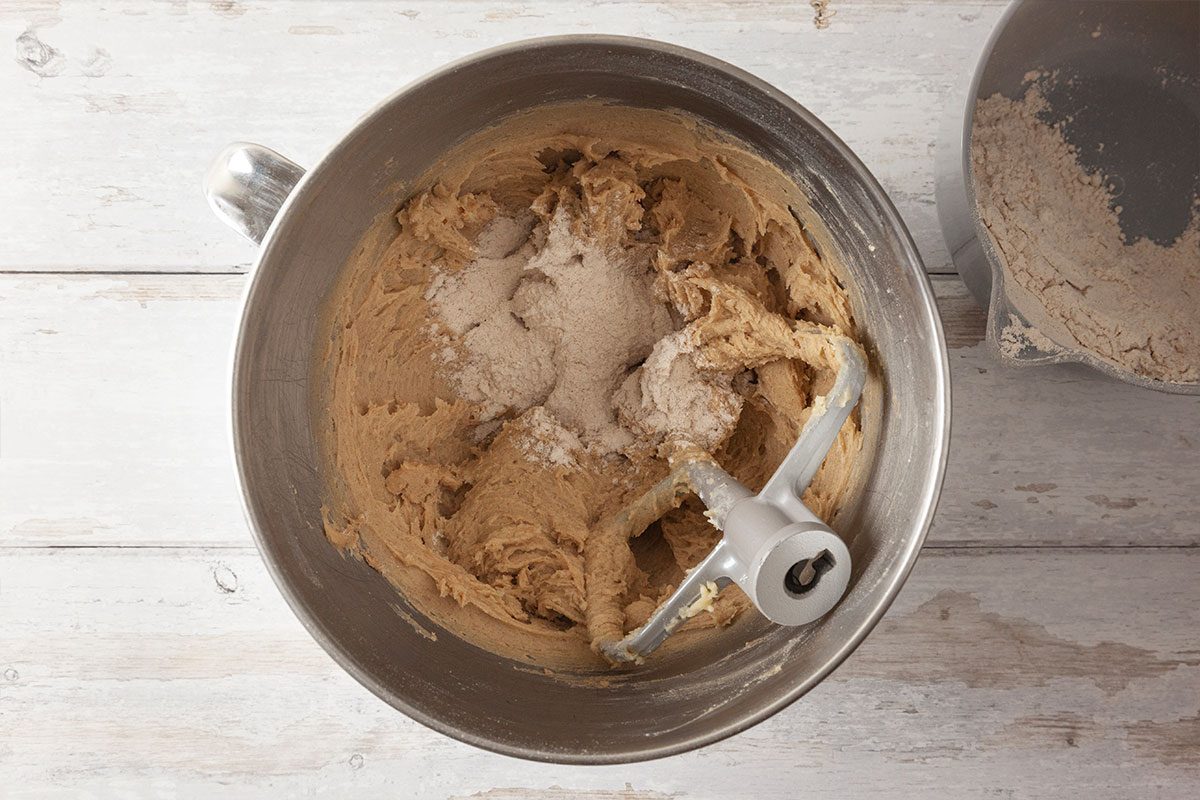 A metal mixing bowl containing cookie dough and flour, with a paddle attachment resting in the bowl, sits on a light wooden surface. Another small bowl with flour is partially visible to the right.