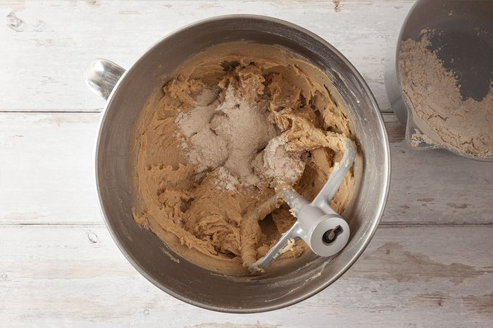 A metal mixing bowl containing cookie dough and flour, with a paddle attachment resting in the bowl, sits on a light wooden surface. Another small bowl with flour is partially visible to the right.