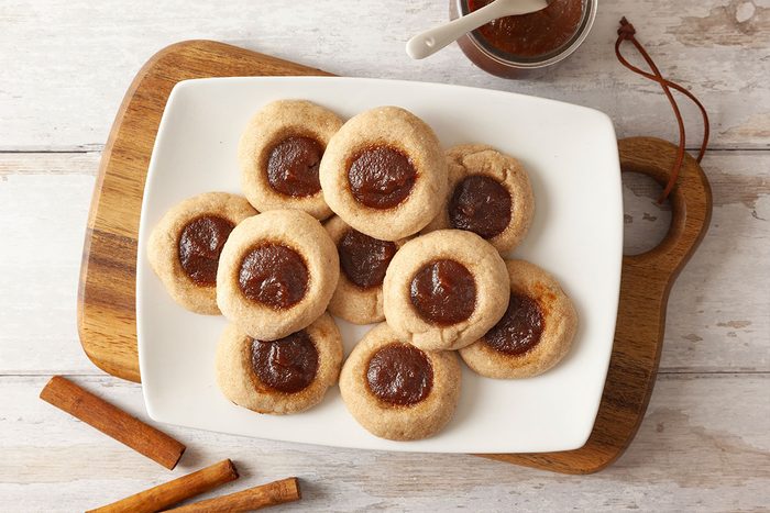 A white plate with round thumbprint cookies filled with brown jam sits on a wooden board. A small jar of jam and two cinnamon sticks are nearby on a light wood surface.