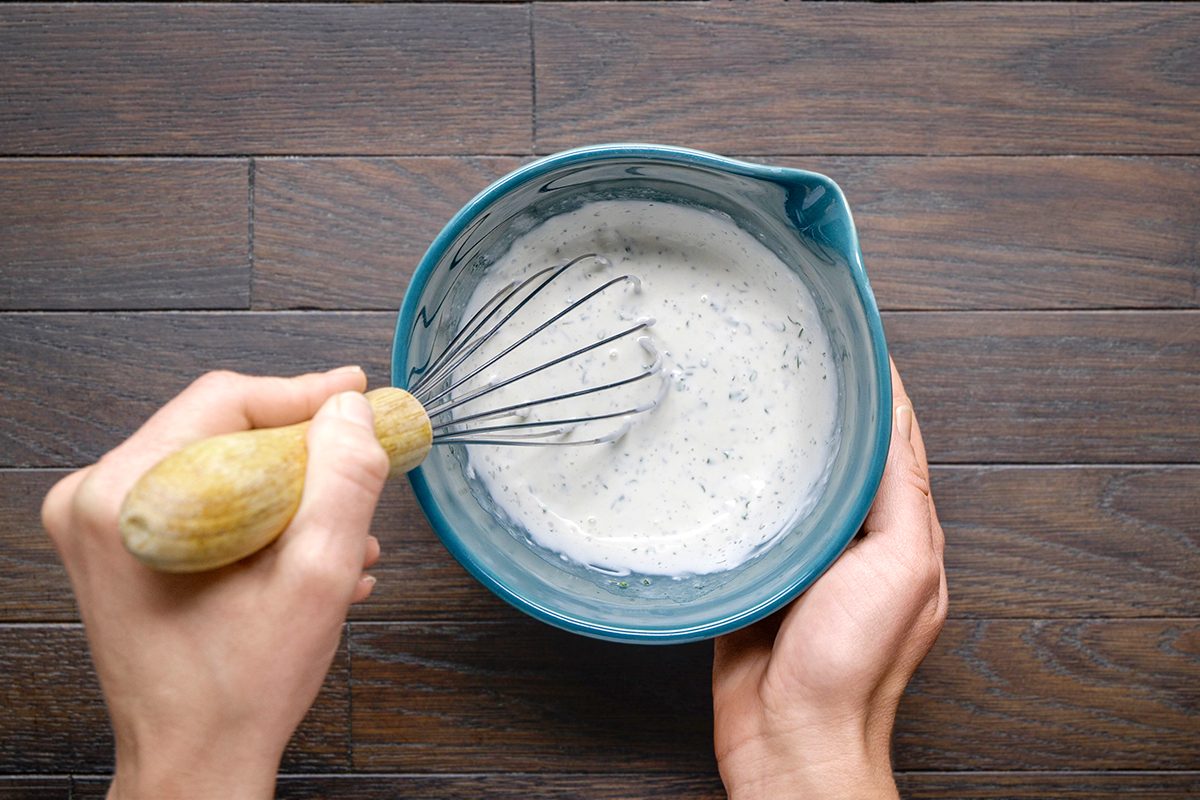 A person holding a blue mixing bowl and whisking a creamy, speckled mixture with a wooden-handled whisk on a dark wooden surface.