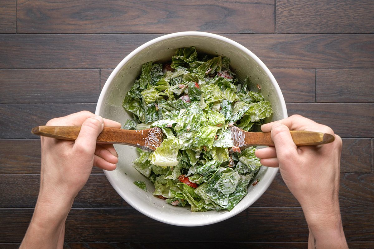 A person tosses a fresh green salad with wooden utensils in a white bowl on a dark wooden table.