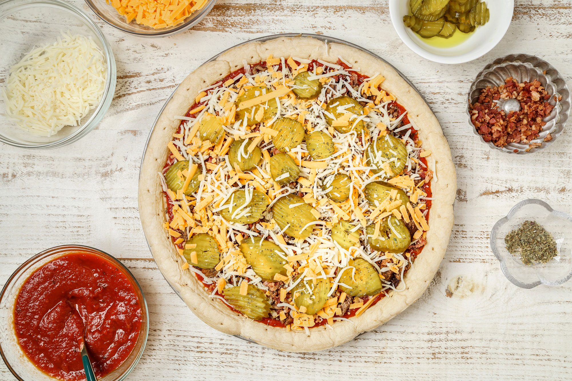Top Shot of a pizza with tomato sauce; shredded cheese; ground meat; and pickles rests on a white wooden surface; surrounded by bowls of cheese; pickles; bacon bits; Italian herbs; and sauce. The setting is bright with a rustic vibe