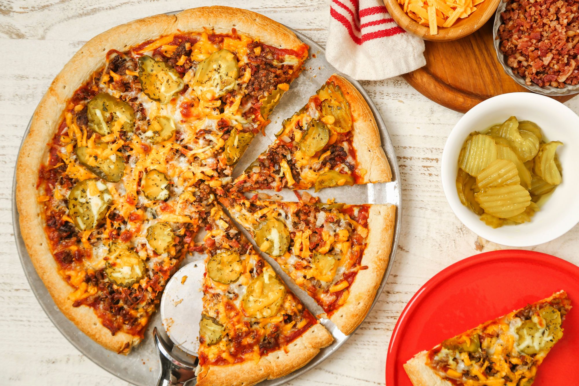 Top Shot of a cheeseburger pizza is sliced on a wooden table; topped with pickles; ground beef; cheese; and onions. Nearby are bowls of pickles; shredded cheese; bacon bits; and a slice of pizza on a red plate