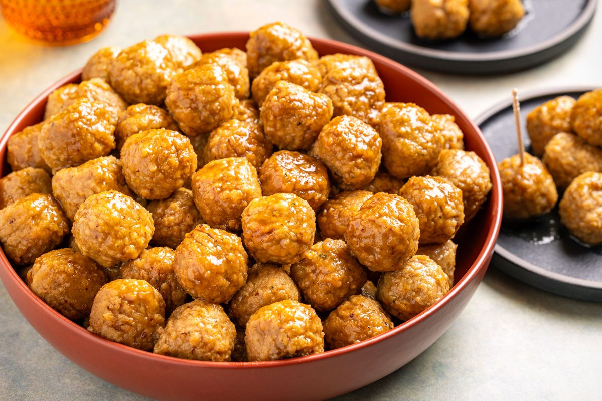 Overhead shot; a large bowl of shiny glazed meatballs; small black plates with extra meatballs in background