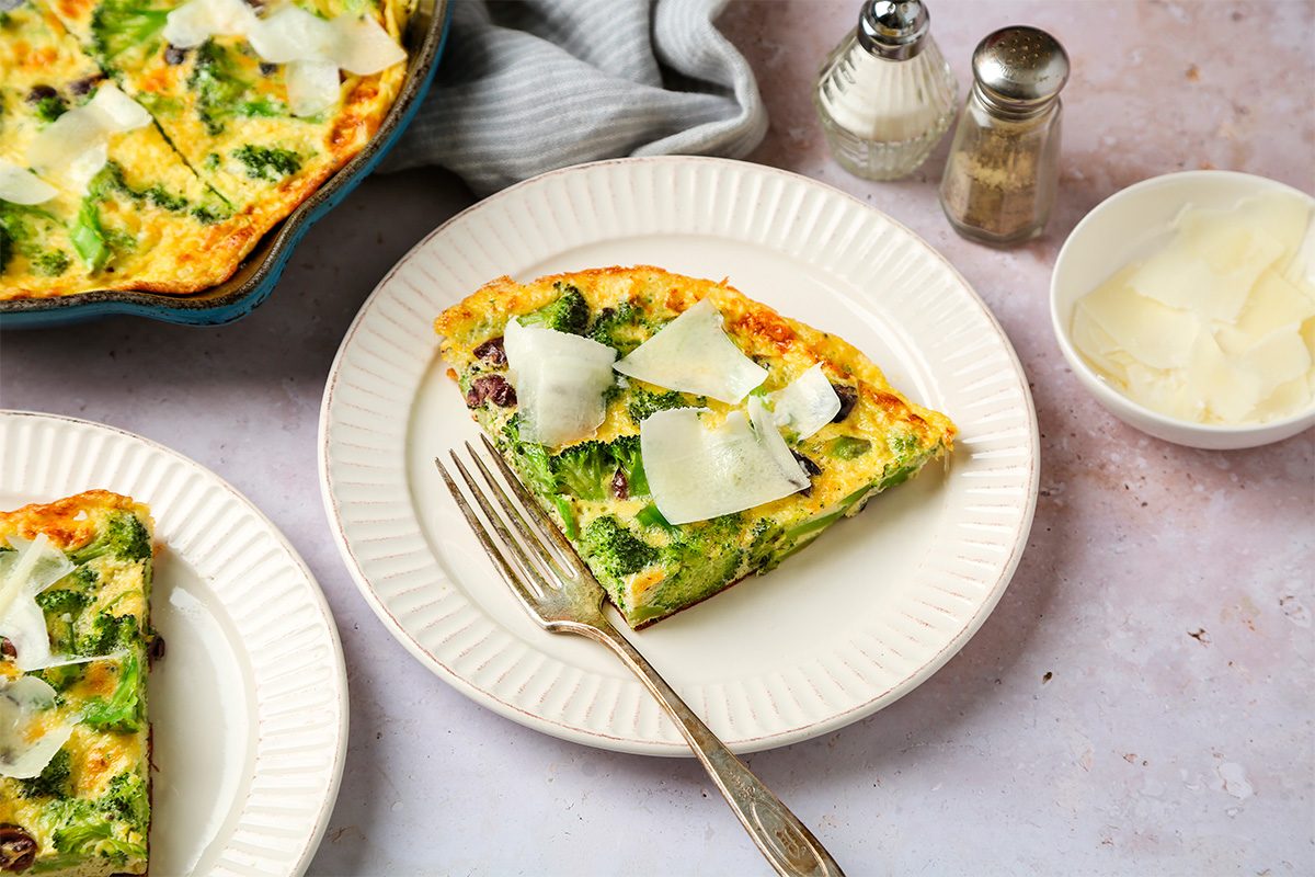 A slice of vegetable frittata topped with cheese shavings on a white plate with a fork, next to a bowl of cheese, salt and pepper shakers, and a striped napkin.
