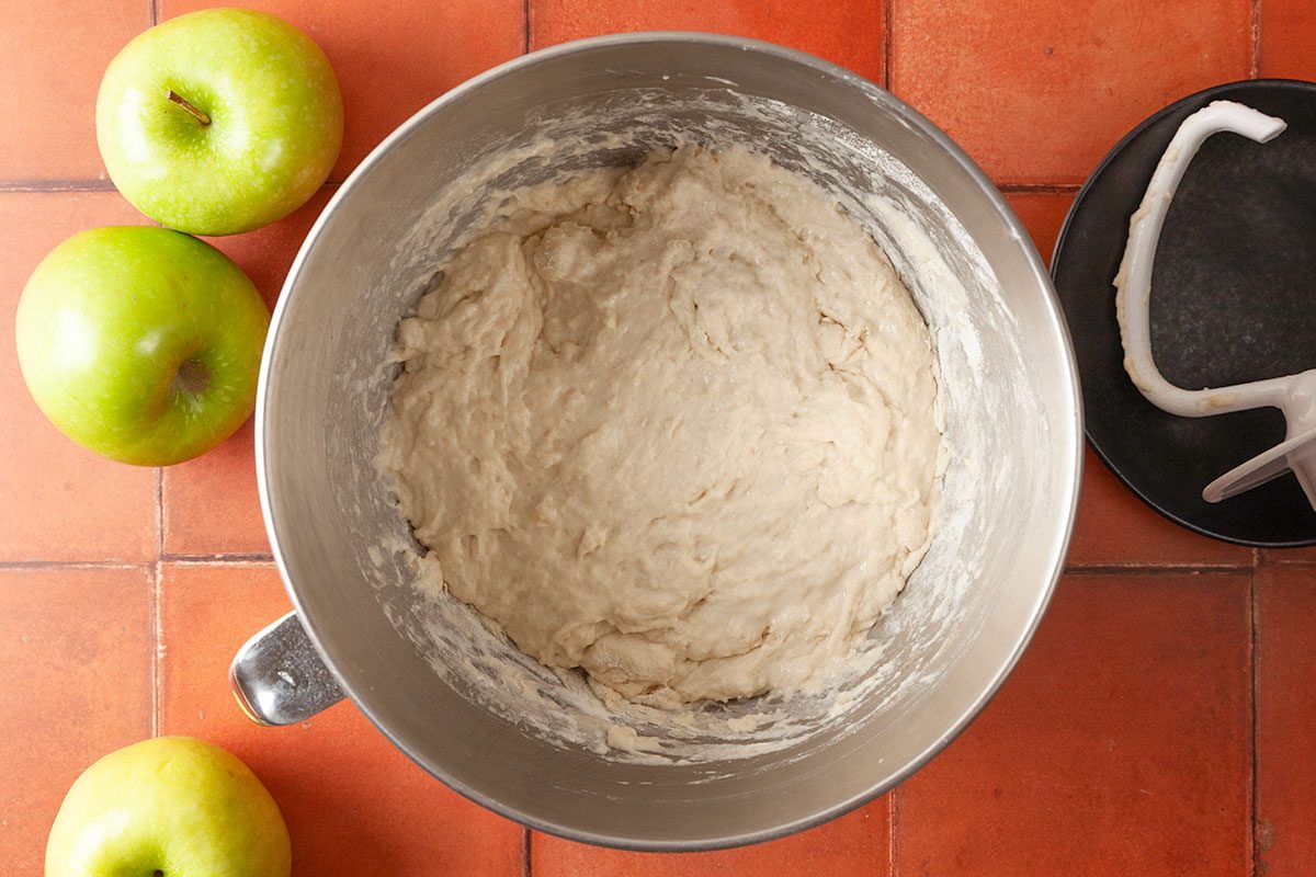 Overhead shot of a metal mixing bowl filled with sticky, unbaked dough, set on a tiled countertop beside three green apples and a black plate holding a dough hook attachment;