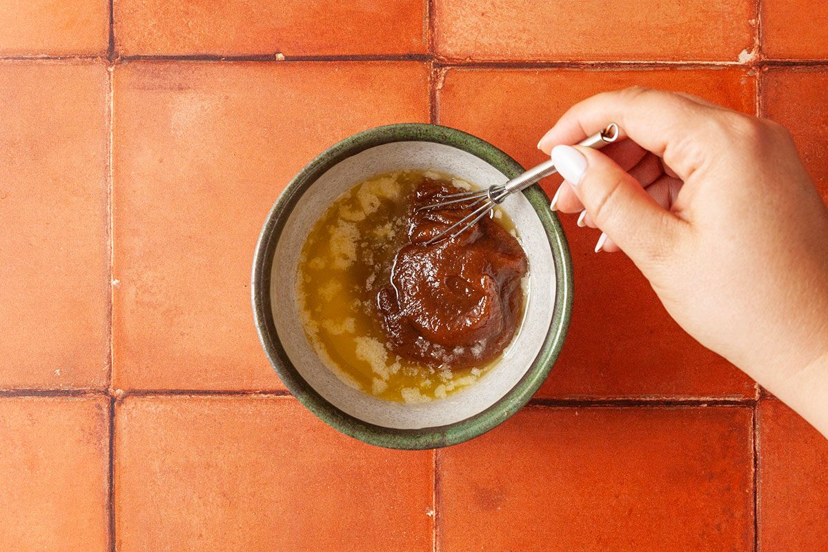 Overhead shot of a hand using a small whisk to mix a brown paste with melted butter in a bowl on a reddish-brown tiled surface.