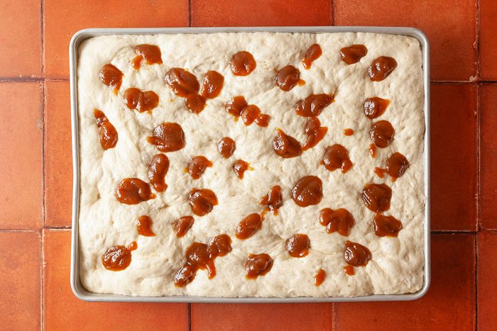 Overhead shot of a rectangular baking pan filled with unbaked dough topped with scattered dollops of brown spread, set on a reddish-brown tiled surface.