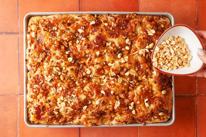 Overhead shot of a hand sprinkling chopped peanuts from a bowl onto a rectangular tray of golden baked bread or cake topped with caramelized crunchy bits; set on a reddish tiled surface.