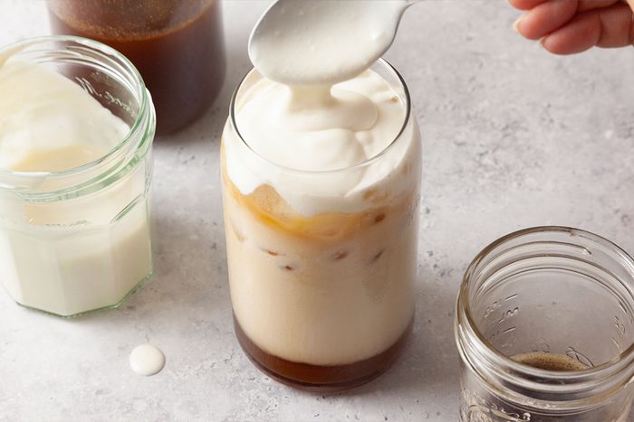 A hand is spooning creamy foam onto a glass of iced coffee. Surrounding the glass are jars containing milk, coffee, and syrup on a light-colored surface.