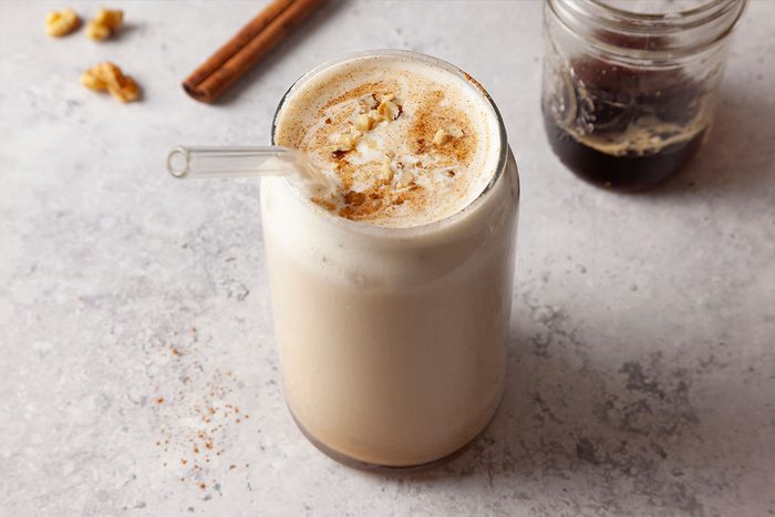 A glass of iced chai latte topped with cinnamon and a glass straw sits on a light surface. A jar of dark liquid and a cinnamon stick are in the background.