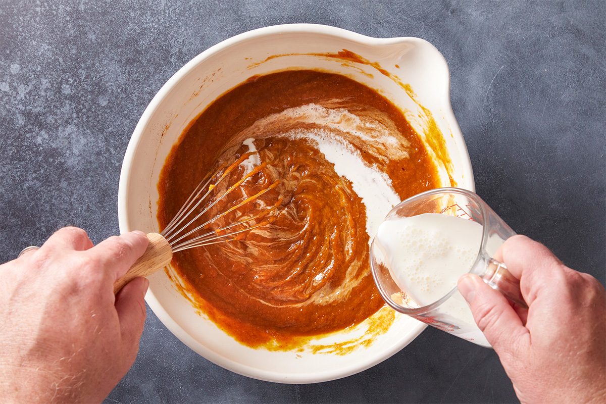 A person whisks a thick orange-brown mixture in a large white bowl while pouring milk from a small glass measuring cup onto the mixture. The background is a dark gray countertop.