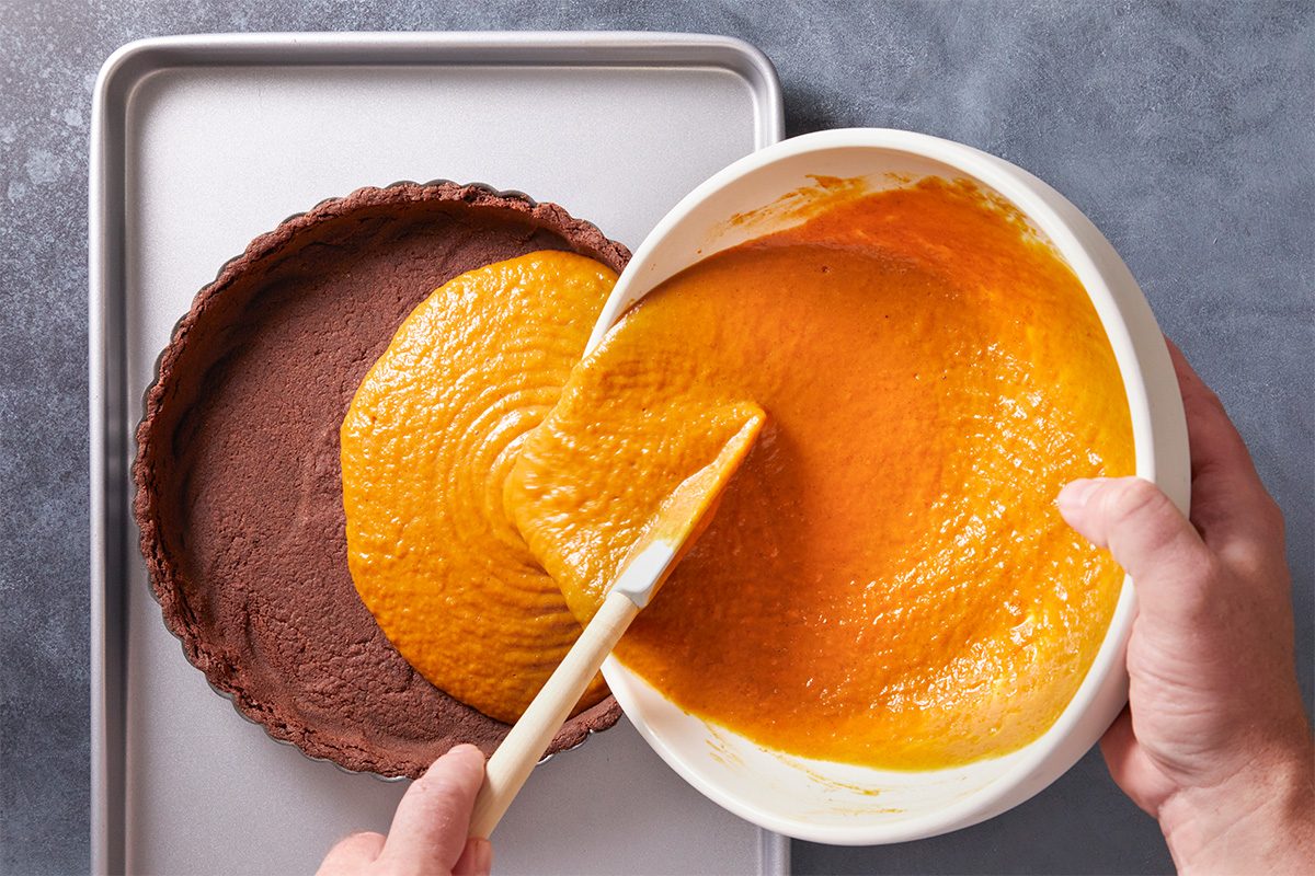 A person pours orange pumpkin pie filling from a white bowl into a chocolate pie crust on a baking sheet, using a spatula to spread the filling evenly.