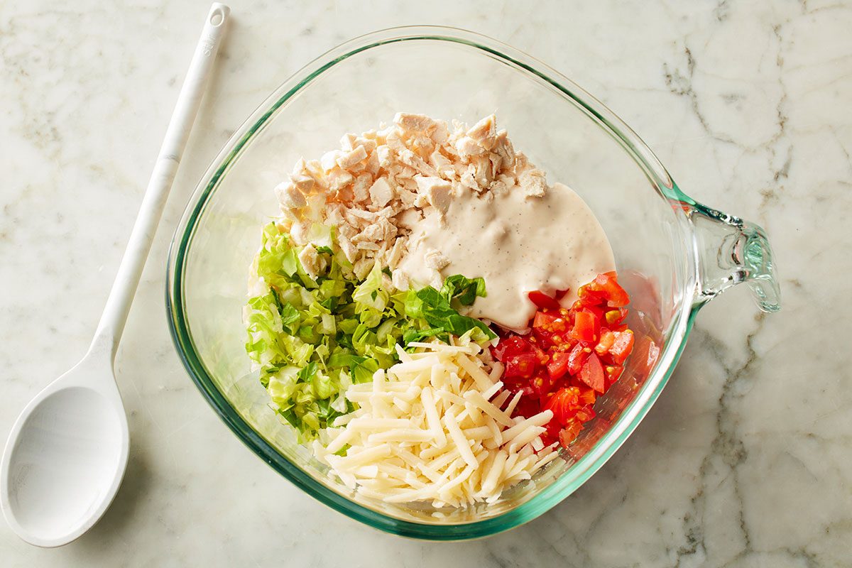Close-up shot of a fresh salad in a glass bowl featuring chopped lettuce; shredded chicken; diced tomatoes; shredded cheese; and creamy dressing on a marble counter with a white spoon beside it
