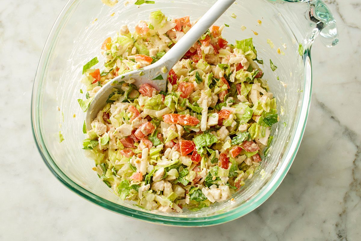 This is a close-up shot of a glass bowl containing freshly chopped salad with lettuce; tomatoes; assorted vegetables; and creamy dressing; accompanied by a white serving spoon on a marble countertop