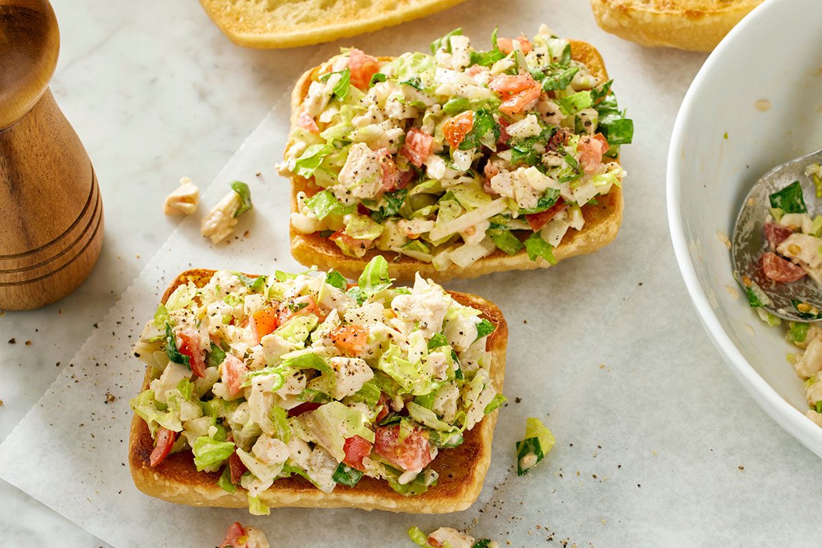 vThis is an overhead shot showing two open-faced chicken salad sandwiches on toasted bread atop parchment; nearby are a bowl of extra salad with a spoon and a pepper grinder on the left