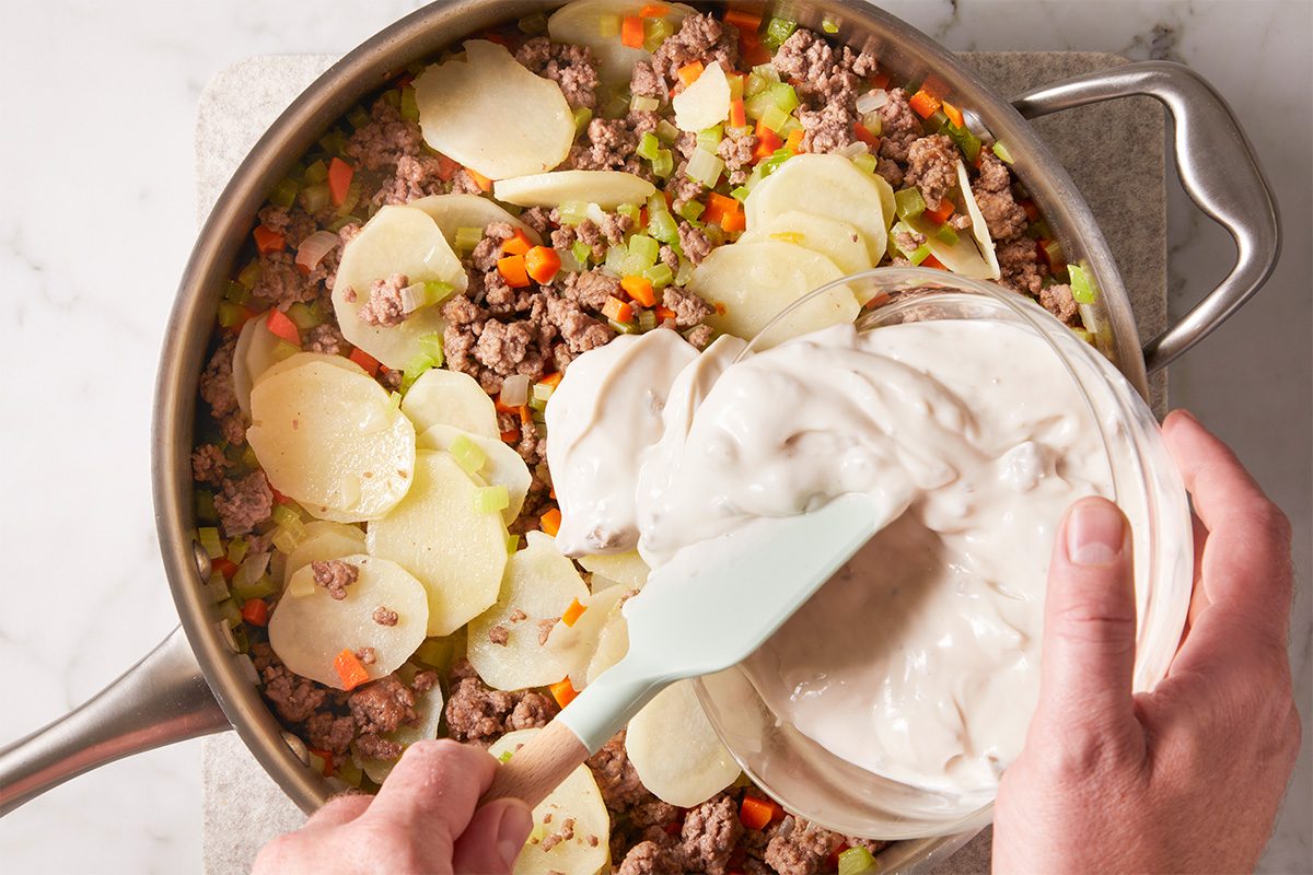 A person spreads creamy sauce over a skillet filled with sliced potatoes, ground meat, chopped carrots, and celery on a marble countertop.