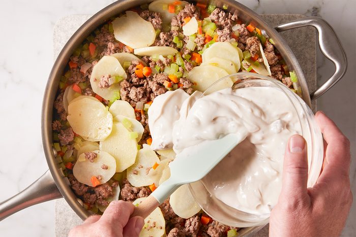 A person spreads creamy sauce over a skillet filled with sliced potatoes, ground meat, chopped carrots, and celery on a marble countertop.