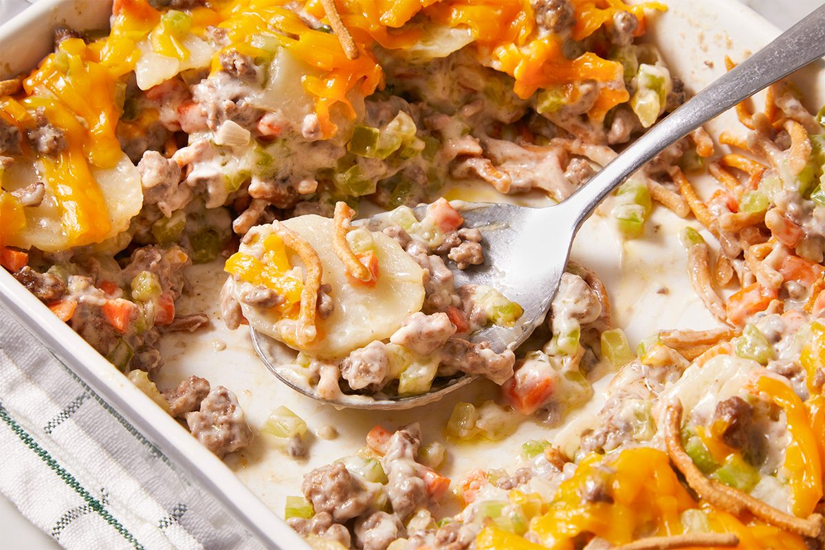 A close-up of a casserole dish filled with a creamy mixture of ground beef, cheese, potatoes, vegetables, and sauce. A metal spoon is scooping out a portion, revealing the hearty ingredients inside.