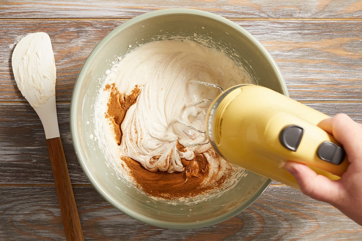 A hand using a yellow electric mixer blends brown and white ingredients in a green mixing bowl on a wooden surface; a spatula with batter rests nearby.