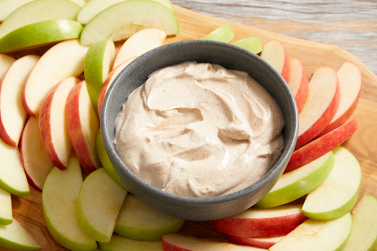 A bowl of creamy brown dip sits on a wooden board surrounded by neatly arranged slices of red and green apples.