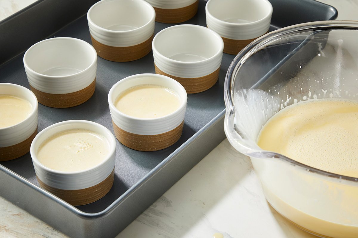 A metal baking pan holds several white and brown ramekins filled with custard mixture. Beside the pan, a clear mixing bowl with remaining custard sits on a white countertop.