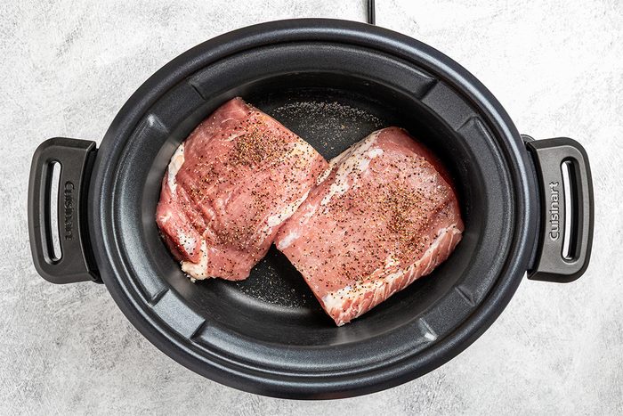 Two raw, seasoned pieces of pork sit side by side in a black slow cooker on a light gray countertop.