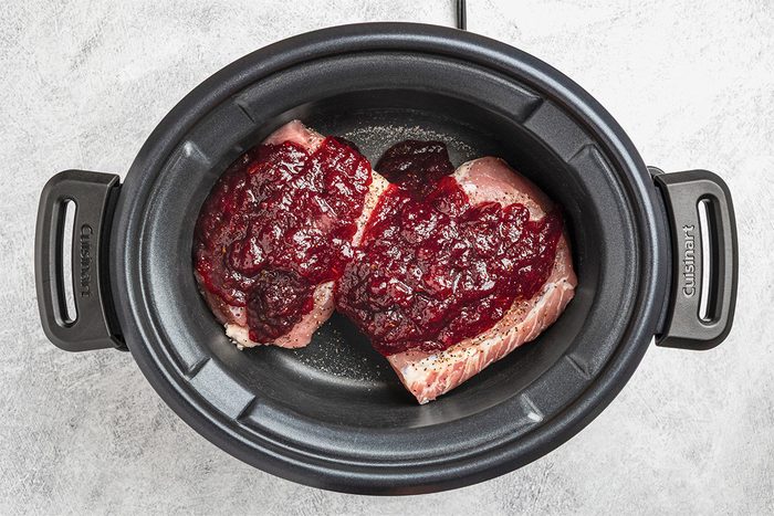 Two pieces of raw meat in a black slow cooker topped with a thick, red sauce, possibly cranberry sauce, on a light gray surface.