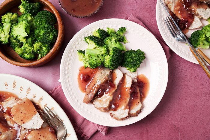 overhead shot of three plates of food on a pink surface, the center plate has pork tenderloin, broccoli florets, and cranberry sauce; the plate to the left has pork tenderloin and a fork; the plate to the right has pork tenderloin and two forks; there is a small bowl of cranberry sauce and a small bowl of broccoli florets on the left