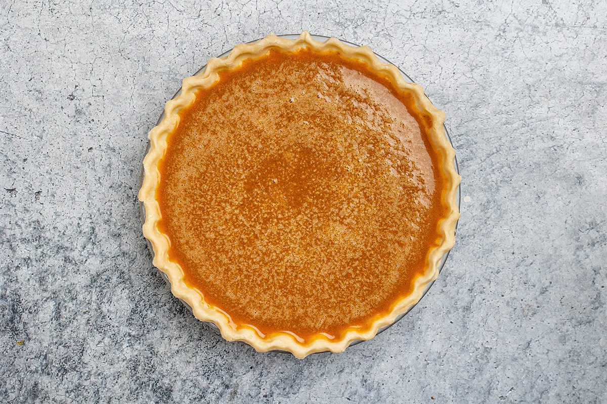 A whole pumpkin pie with a golden, slightly glossy filling and crimped crust sits on a textured gray surface, viewed from above.