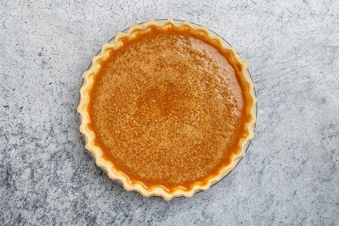 A whole pumpkin pie with a golden, slightly glossy filling and crimped crust sits on a textured gray surface, viewed from above.