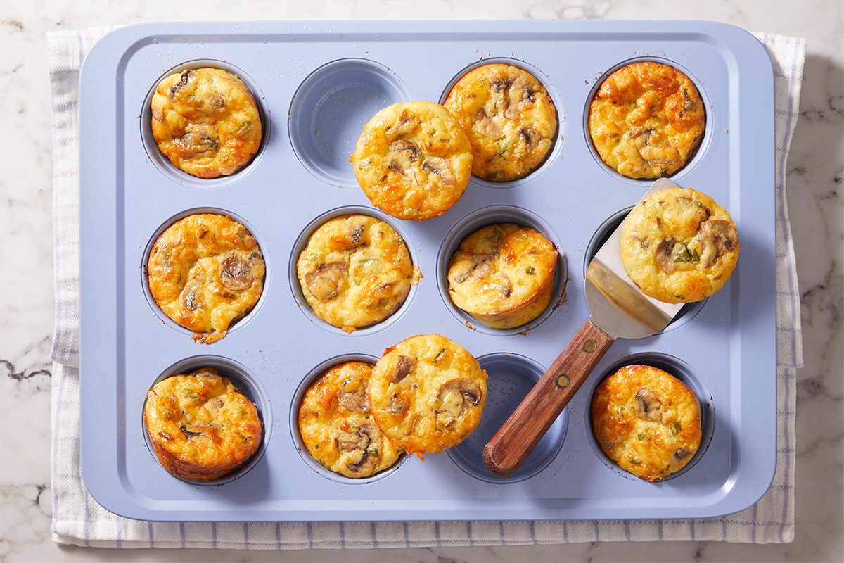 A blue muffin tin holds eleven baked egg muffins with visible pieces of vegetables and cheese; one muffin is being lifted with a spatula. The tin rests on a striped cloth on a marble surface.