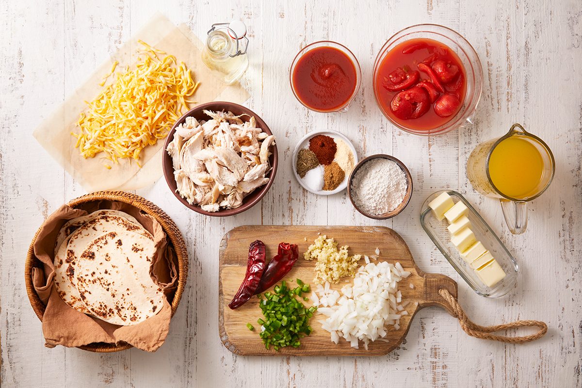 Ingredients for a recipe are arranged on a white surface. They include shredded cheese, shredded chicken, tomato sauce, chopped tomatoes, chicken broth, seasonings, flour, chopped onion, garlic, green pepper, tortillas, dried chilis, and butter.