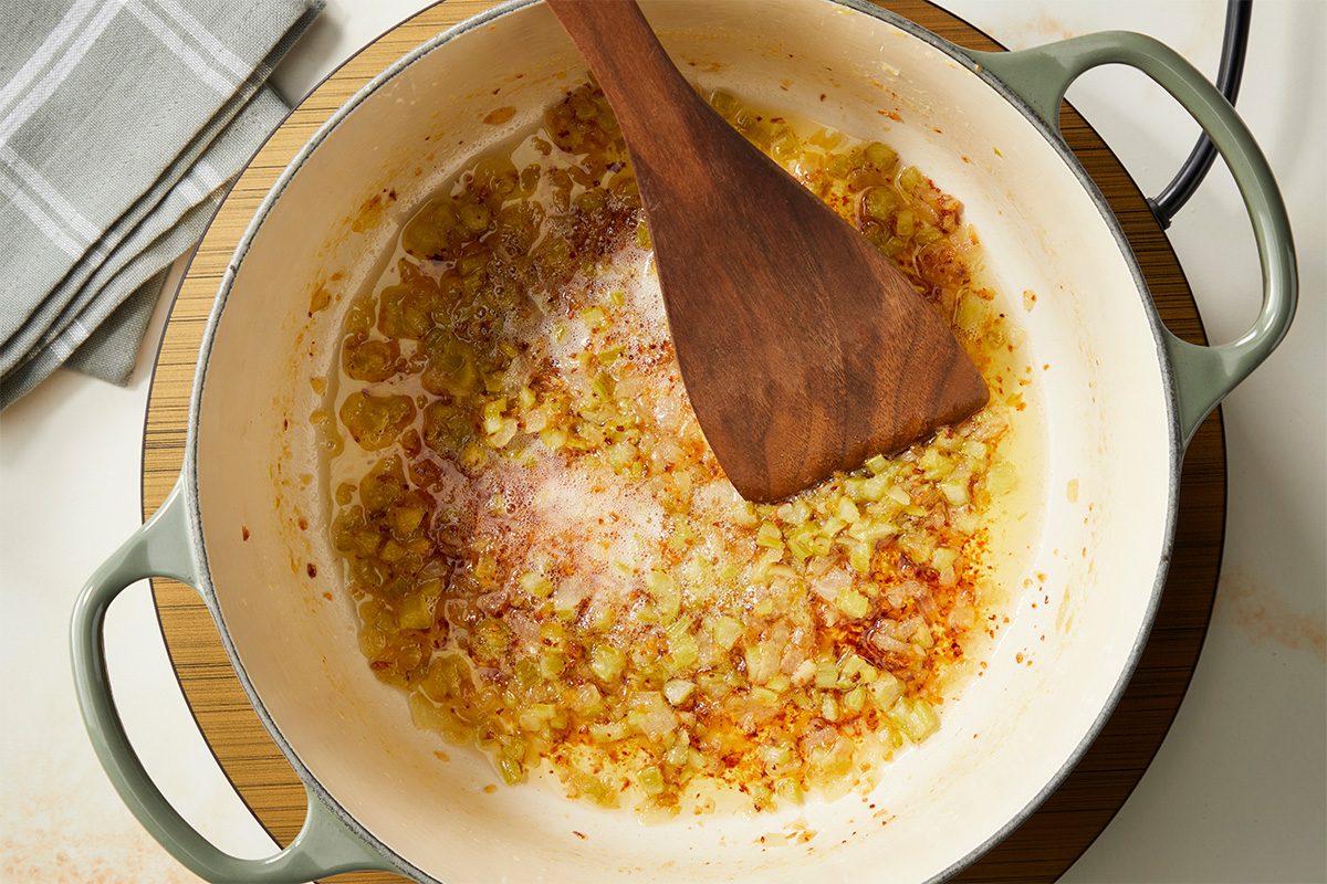Chopped onions sautéing and turning golden brown in a light-colored pot, stirred with a wooden spatula. A folded gray-striped kitchen towel is nearby.