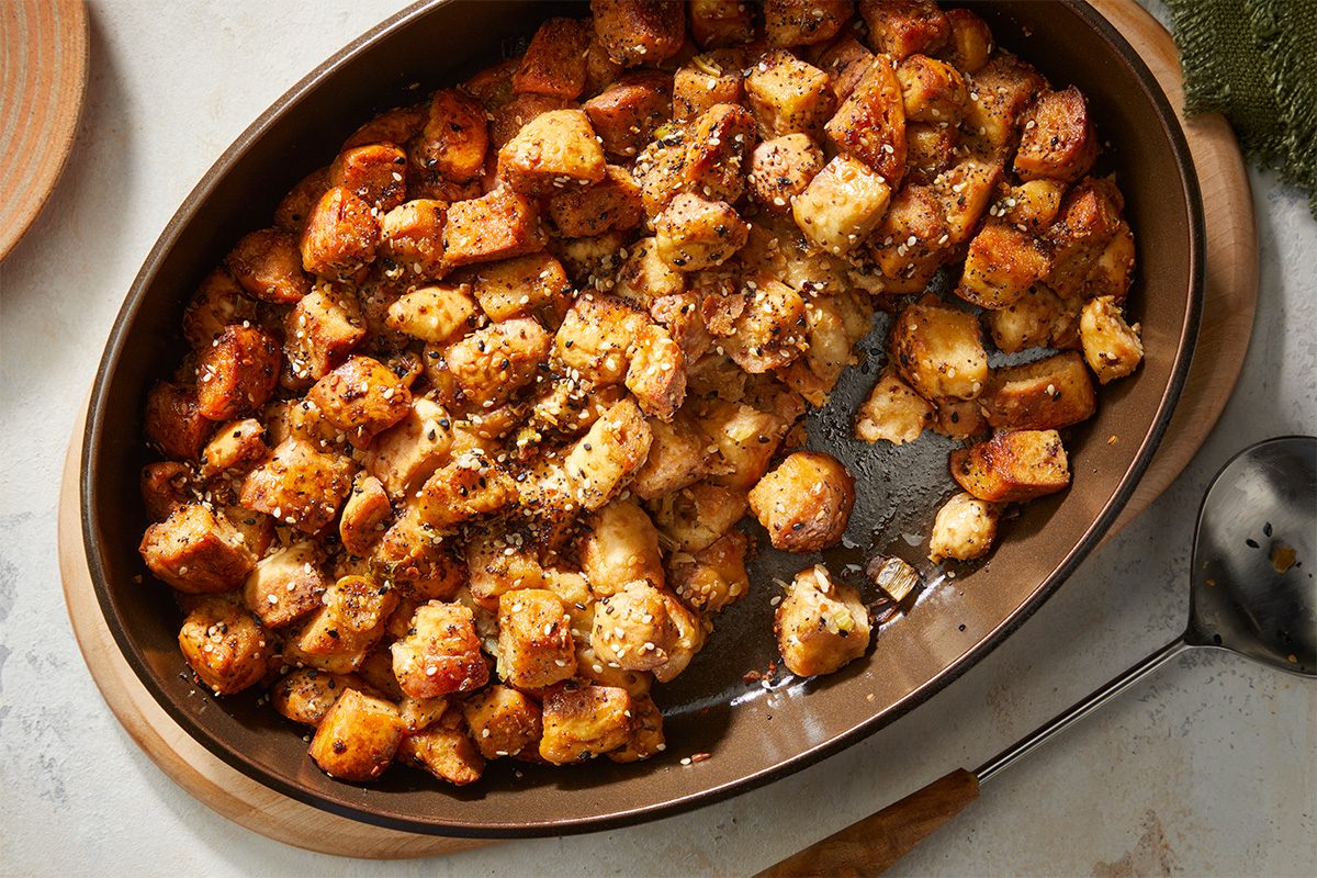 A brown oval baking dish filled with golden-brown, seasoned bread cubes, resembling baked stuffing or croutons, sits on a light countertop next to a serving spoon.