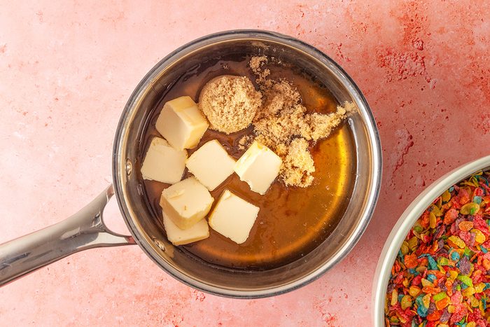 Overhead shot of a saucepan with cubed butter and brown sugar on a pink surface, next to a bowl of colorful cereal pieces