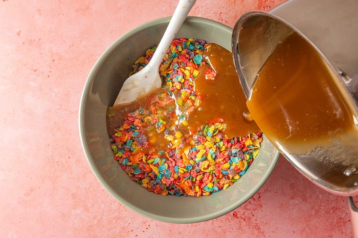 Overhead shot of a bowl of colorful Fruity Pebbles cereal being mixed with caramel sauce from a saucepan, using a white spatula, on a pink surface