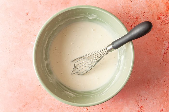Overhead shot of a green bowl filled with smooth white batter, being whisked with a black-handled metal whisk, set on a light pink countertop