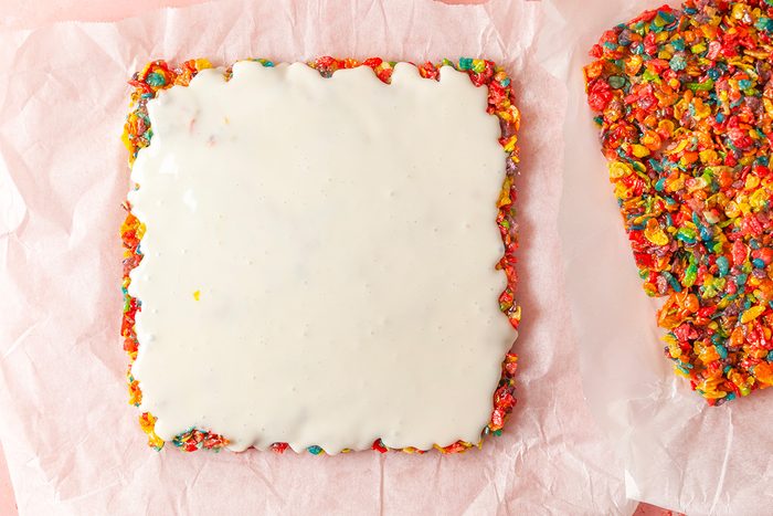 Overhead shot of a square colorful cereal dessert topped with smooth white icing; set on pink parchment paper, with another piece partially visible beside it