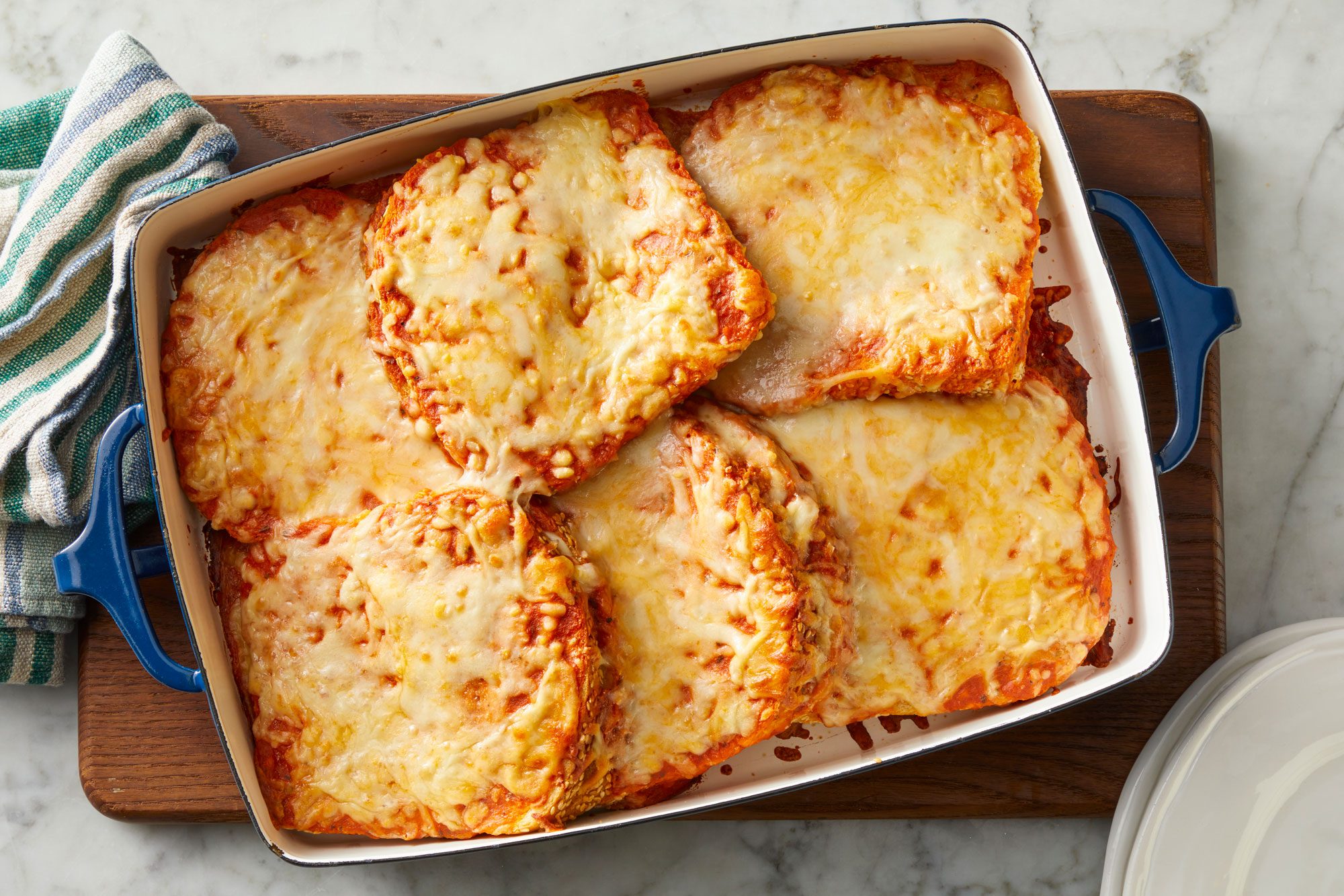 Overhead shot of Grilled Cheese & Tomato Soup Bake in a baking sheet over wooden board with a napkin and empty plates nearby; all set on a marble surface;