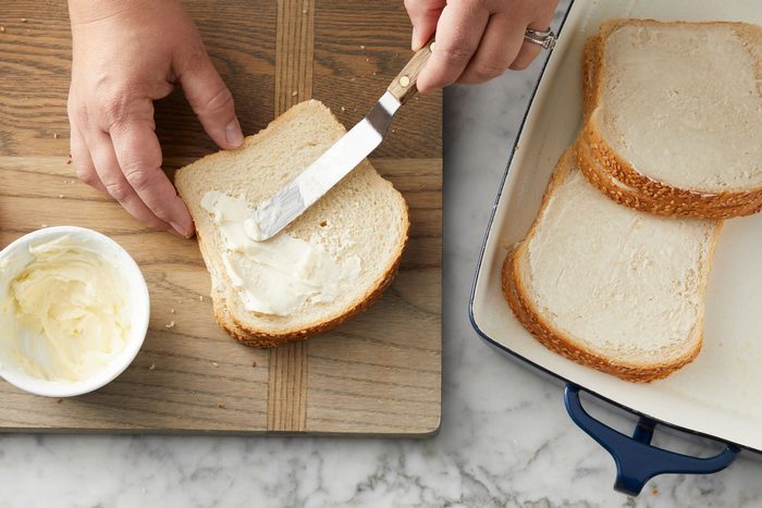 Overhead shot of spread outsides of sandwiches with butter on a wooden tray; arrange in a greased baking dish over a marble surface