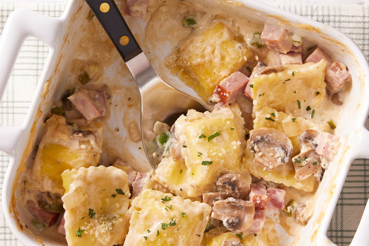 A casserole dish filled with cheesy ravioli, chunks of ham, sliced mushrooms, and creamy sauce, garnished with chopped herbs, with a serving spatula resting inside.