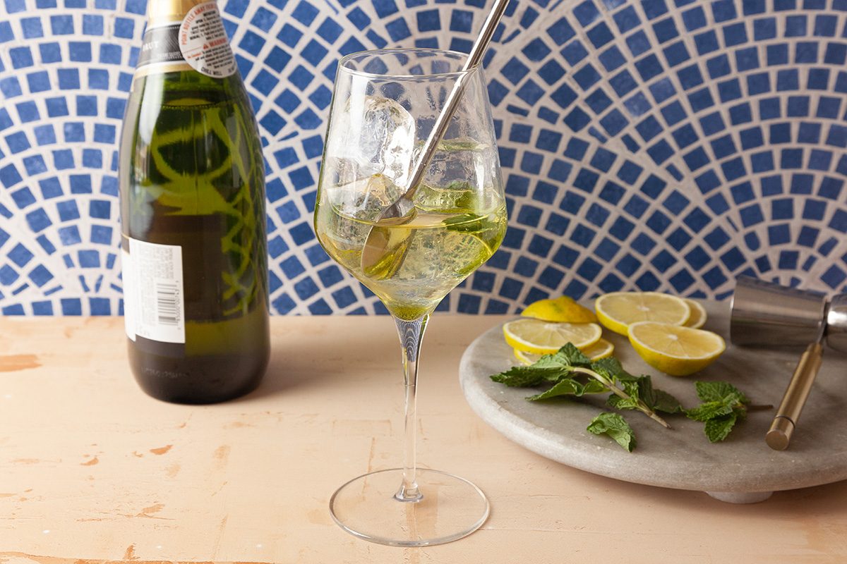 A glass of cocktail with mint leaves and a straw stands on a wooden surface beside a bottle, with sliced lemons, mint, and a jigger on a marble tray; blue patterned tiles are in the background.
