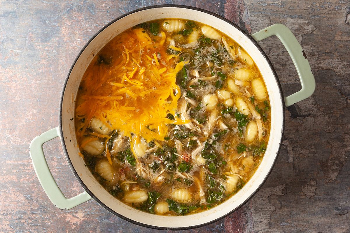 A pot of soup with gnocchi, shredded cheese, leafy greens, and herbs in a light broth, sitting on a rustic surface.