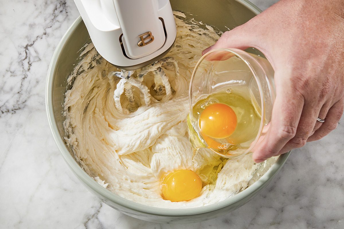 A hand adds a glass bowl with an egg to a mixing bowl containing batter and another egg, while an electric mixer rests above the mixture on a marble countertop.